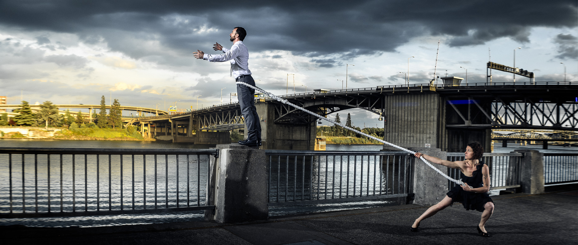 In this scene, Modern dancers Noel Plemons and Rachael Lembo practice social distance in this award-winning image next to the Morrison Bridge. Noel is suspended by a rope and reaches towards the opposite bank of the Willamette River and Rachael keeps the tension on the the opposite end of the rope to stop Noel from falling in. By Andy Batt