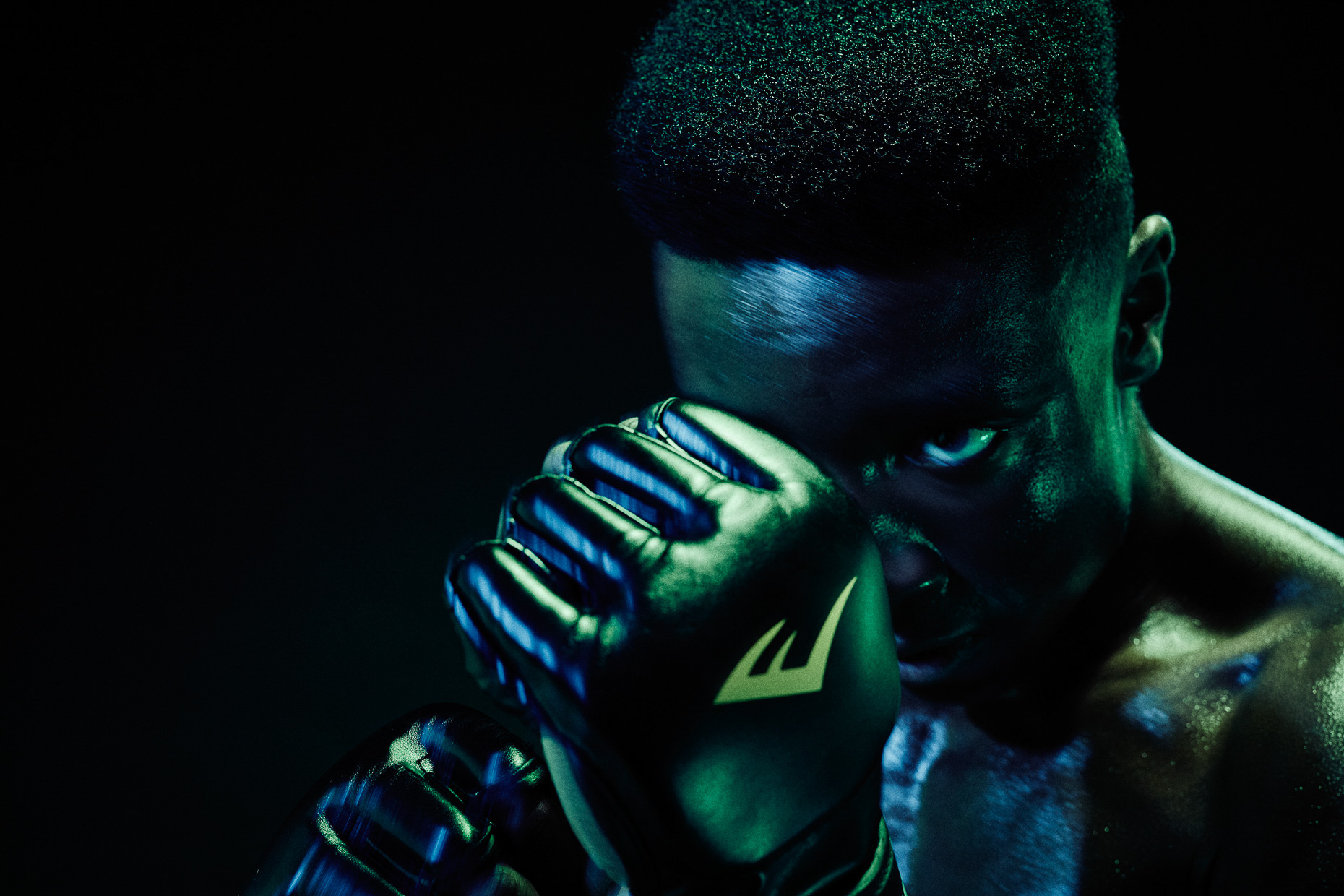Nike boxing MMA Versace Sonachi guarding his face with boxing gloves in a dark studio lit with green and blue gels by Andy Batt
